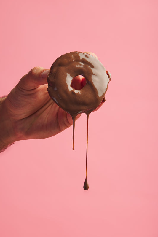 Hand holding a chocolate donut with icing dripping, in front of a pink background